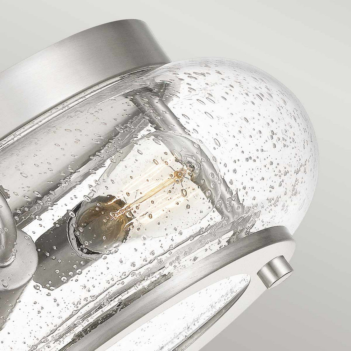 Close-up of the Trilogy 2 Light Flush - Brushed Nickel, featuring clear, bubble-textured seeded glass and a visible Edison bulb within. The brushed nickel finish complements its sleek metallic design, offering a contemporary appearance.