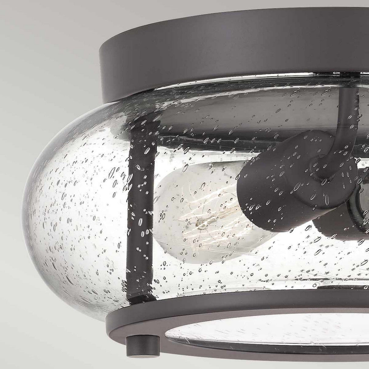 Introducing the Trilogy 2 Light Flush in Old Bronze, featuring a close-up view of its sleek ceiling light fixture with a black metal frame and a clear, seeded glass shade. Inside, the exposed light bulbs are prominently displayed. This design seamlessly blends industrial and contemporary styles with a touch of vintage charm, making it an elegant statement piece for any space.