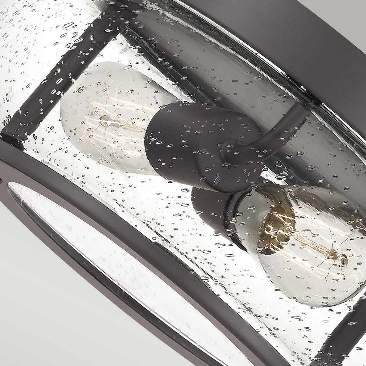 Close-up of the Trilogy 2 Light Flush ceiling light fixture with two exposed bulbs encased in a seeded glass shade, exuding vintage allure. The frame boasts an old bronze color, while the background maintains a neutral grey tone.