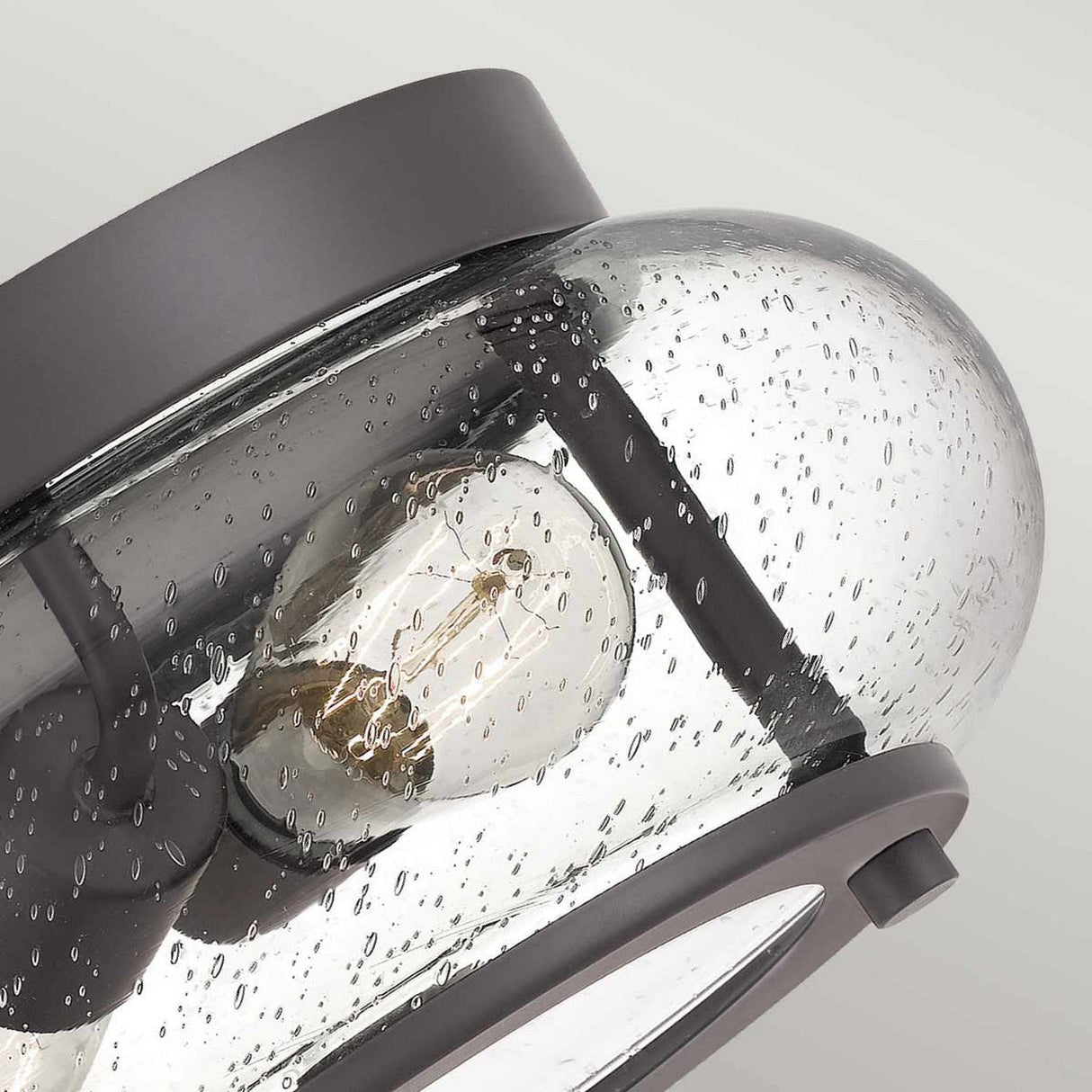 A close-up of the Trilogy 2 Light Flush - Old Bronze features a clear, bubble-textured glass shade. Inside, a visible filament bulb is held by dark metal fixtures finished in old bronze. The background showcases a soft, neutral gray, enhancing the vintage allure of this piece.