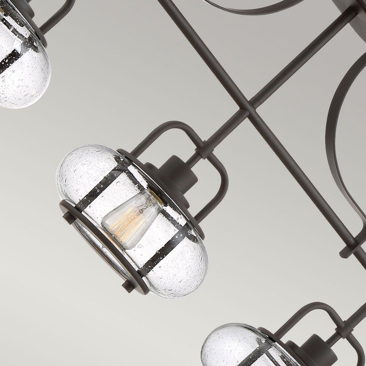 Close-up of the Trilogy 3 Light Island Light in an industrial style, featuring glass pendant shades with visible LED filament bulbs. The fixtures include a metallic frame finished in Olde Bronze and showcase speckled textured glass. The background displays a neutral gradient.