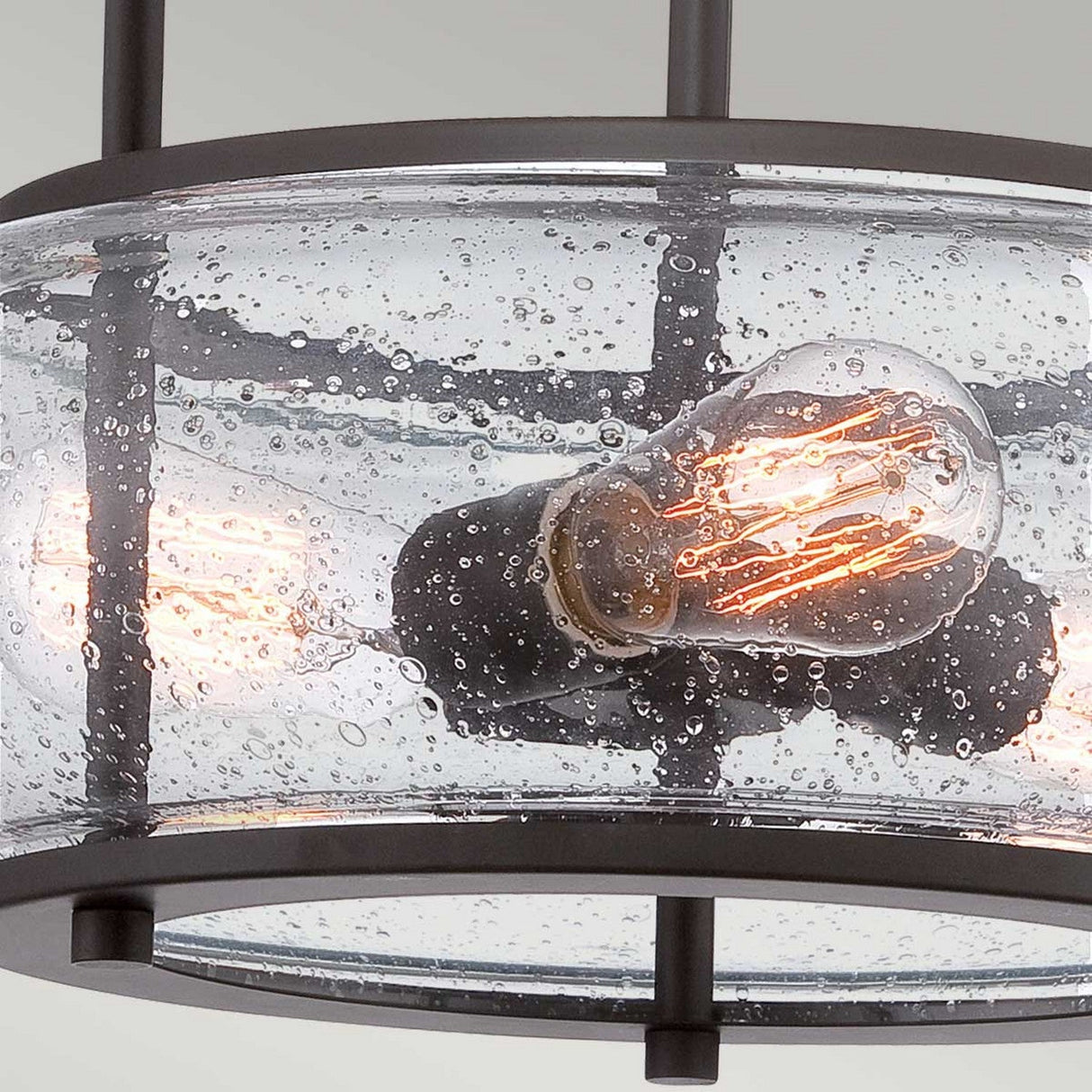 Close-up of the Trilogy 3 Light Medium Semi-Flush in Old Bronze, featuring a modern light fixture with a round, seeded glass enclosure dotted with small bubbles. Inside, exposed filament bulbs emit a warm glow. The fixture's industrial lighting aesthetic is emphasized by its black metal frame and cylindrical design.