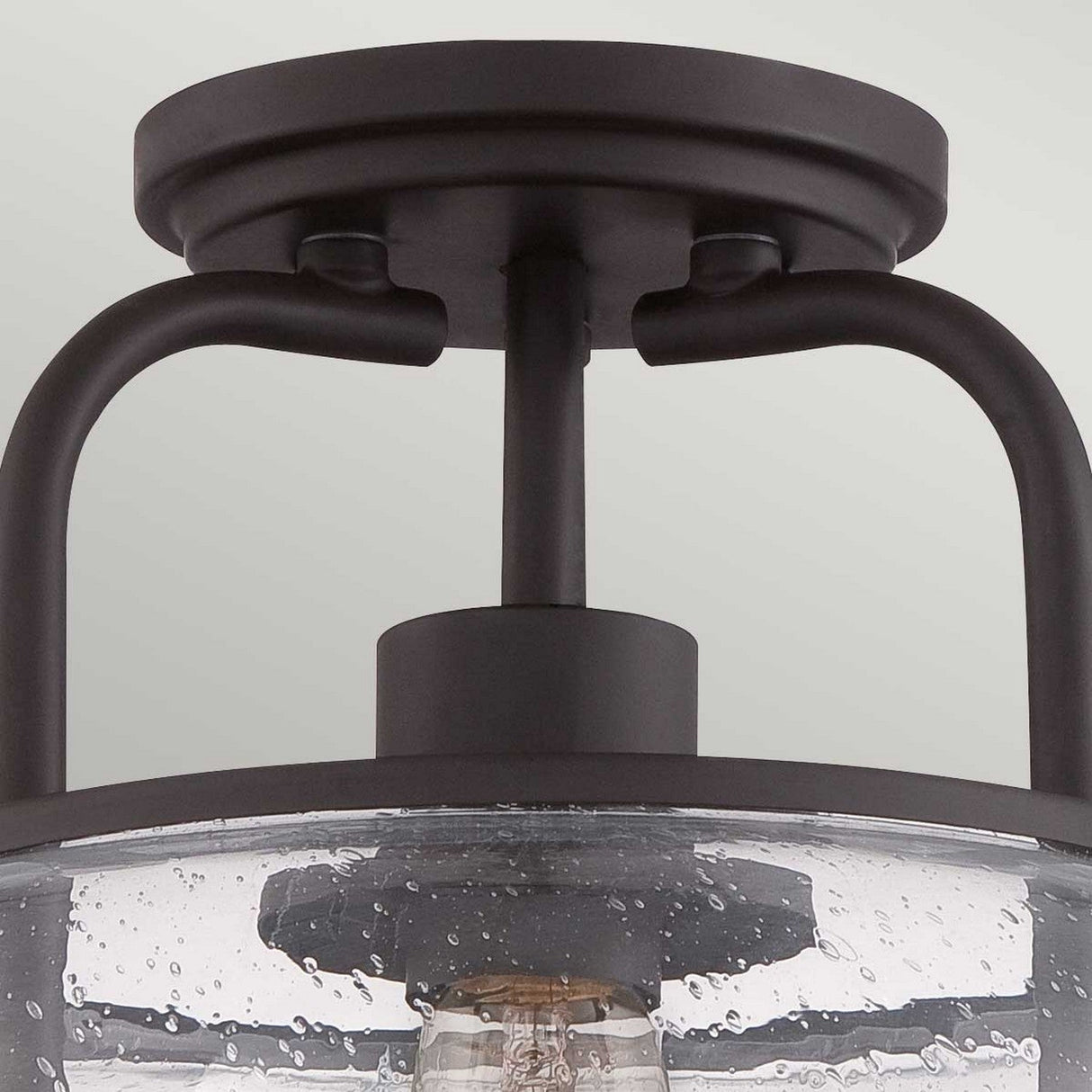 Close-up of the Trilogy 1 Light Small Semi-Flush in Old Bronze, featuring a dark metal frame and a seeded glass shade. This fixture embodies a modern, industrial design with an Edison-style LED filament bulb visible through the textured glass.