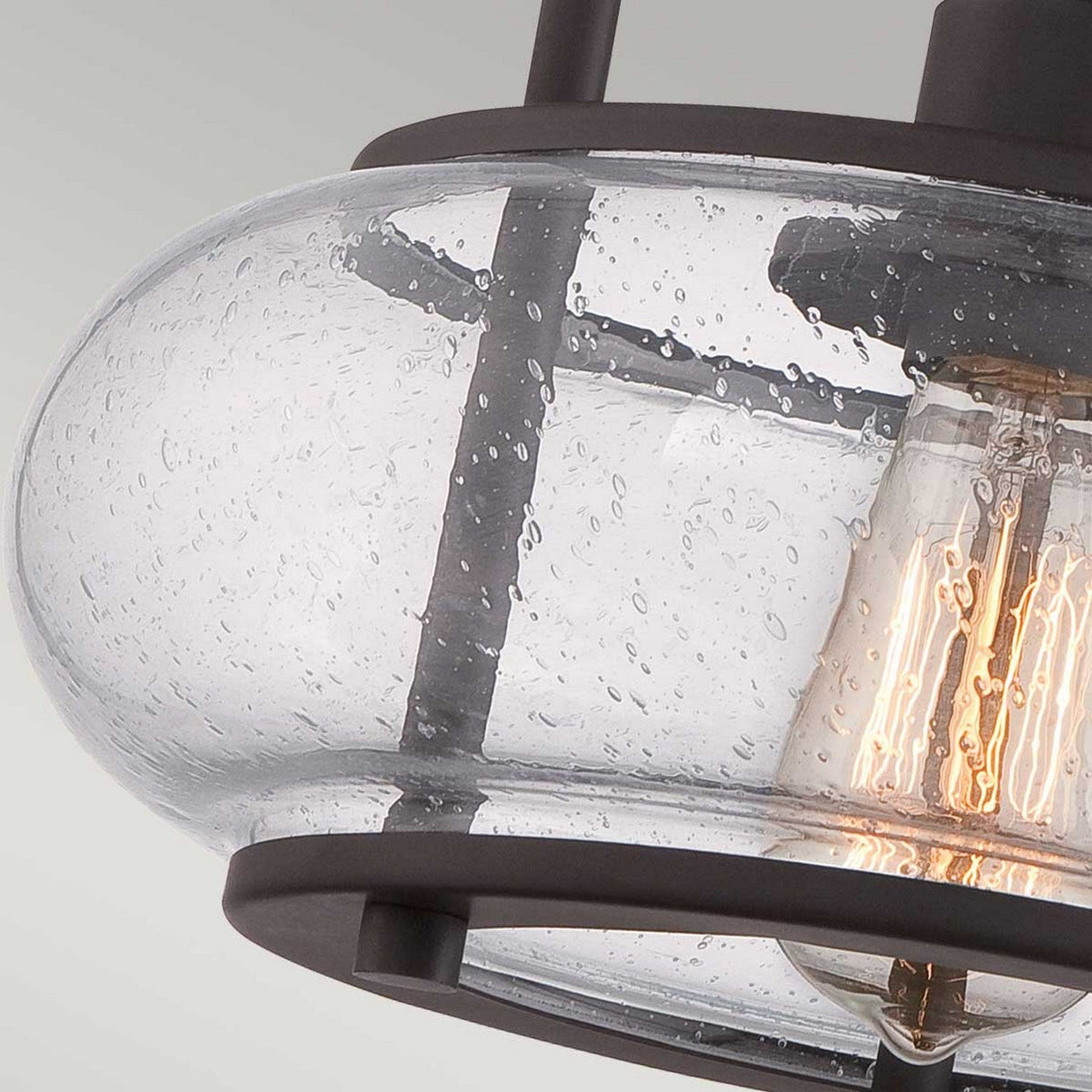 Close-up of the Trilogy 1 Light Small Semi-Flush in Old Bronze, featuring a clear seeded glass shade. Inside, an Edison-style LED filament bulb emits a warm glow, while the Old Bronze finish provides a striking contrast with the textured glass.