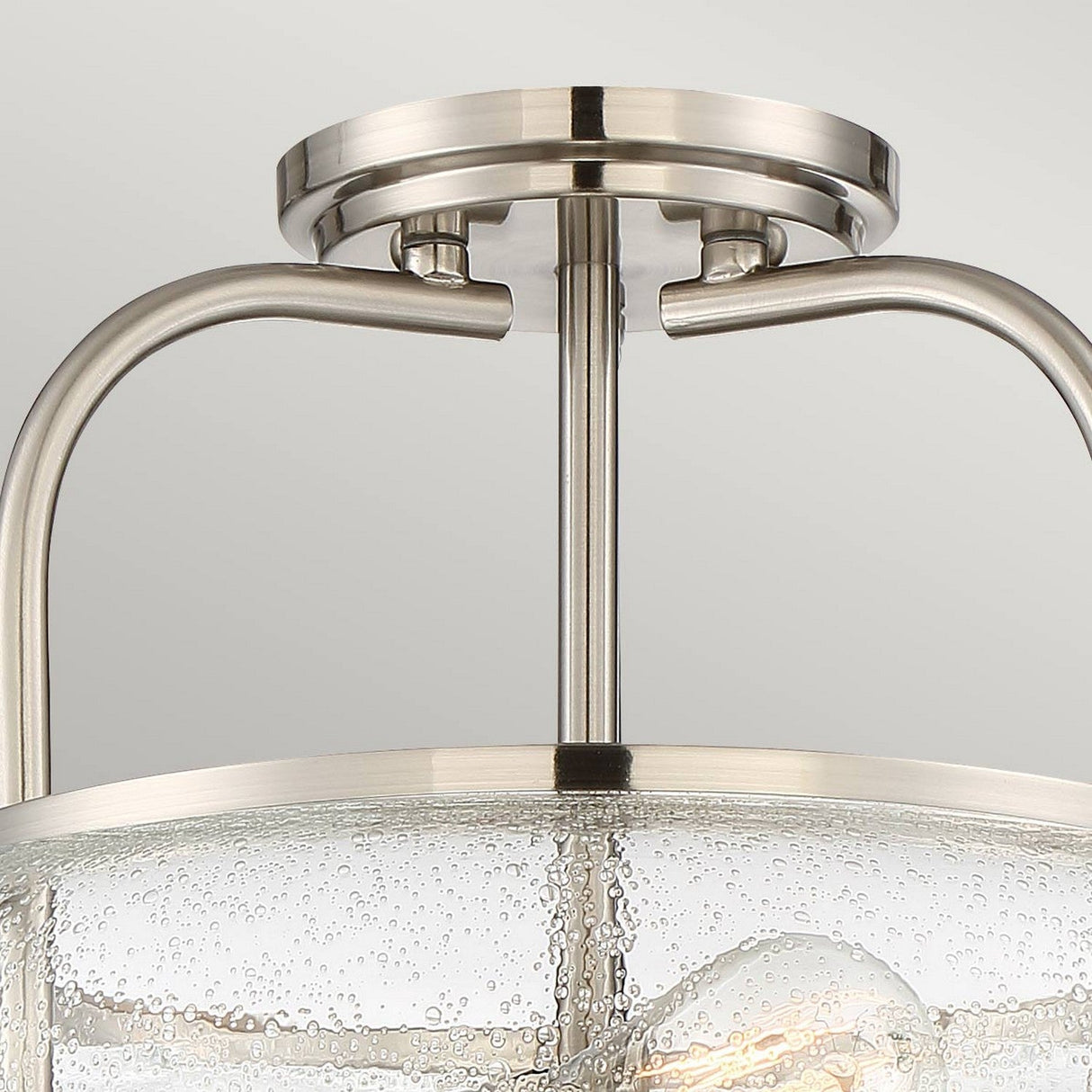 Close-up of the Trilogy 3 Light Semi-Flush Fixture in Brushed Nickel, featuring curved metal arms and a cylindrical clear, seeded glass shade. The shade is adorned with small, bubble-like details that add texture to the design.