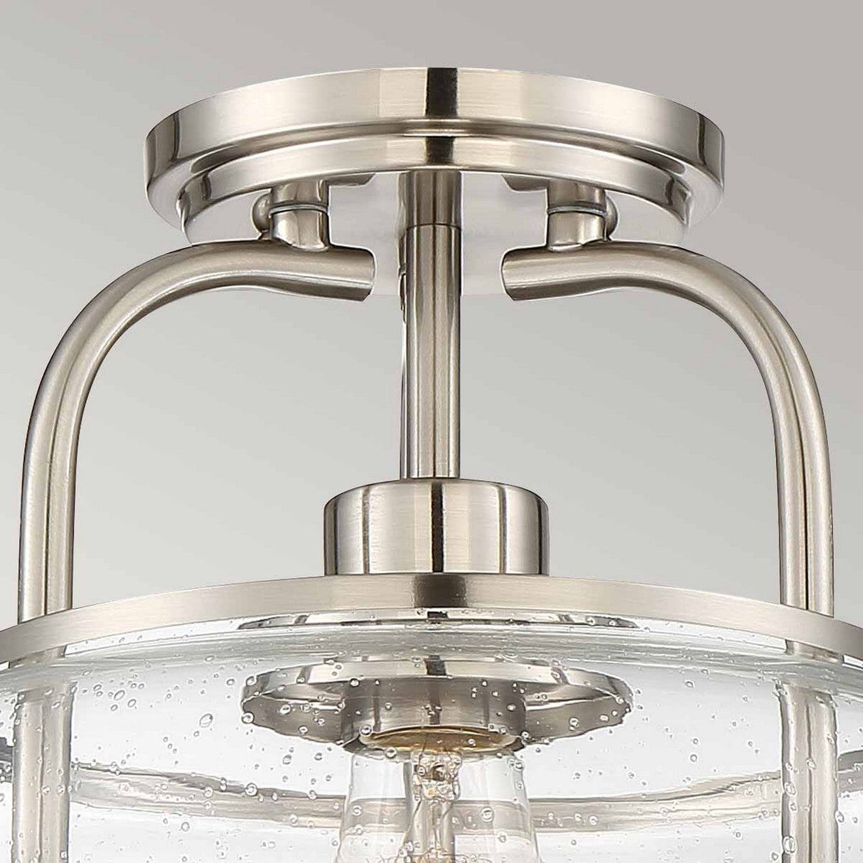 The Trilogy 1 Light Semi-Flush in Brushed Nickel is a ceiling-mounted fixture that boasts a clear seeded glass dome with small bubbles. Its sleek, modern design includes two supporting arms and features an exposed Edison-style LED filament bulb.