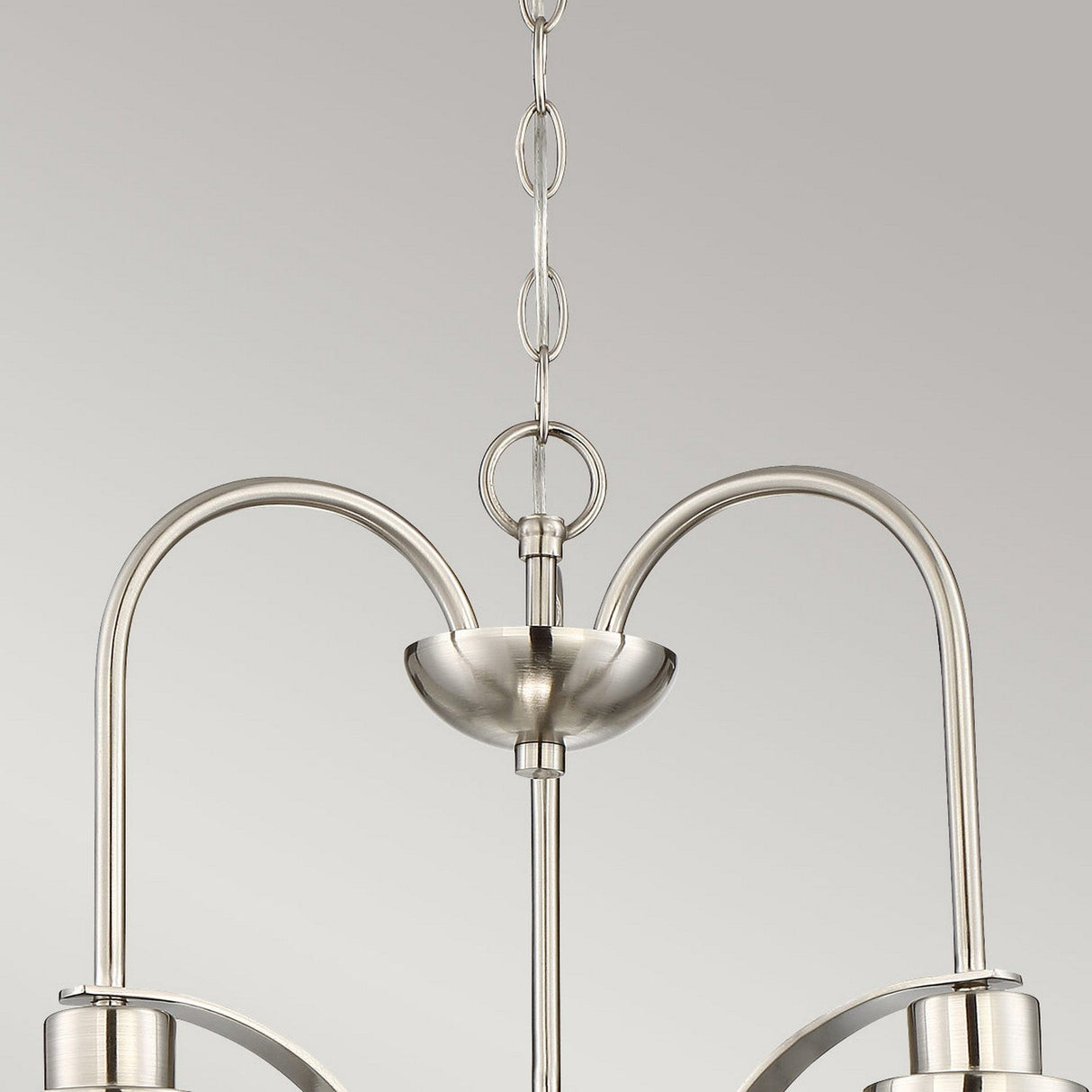 Against a plain gray background, the Trilogy 3 Light Chandelier - Brushed Nickel is showcased up close, featuring its sleek brushed nickel finish and gracefully curved arms hanging from a chain. Its semi-flush design exudes modernity and sophistication, enhancing the elegance of any room.