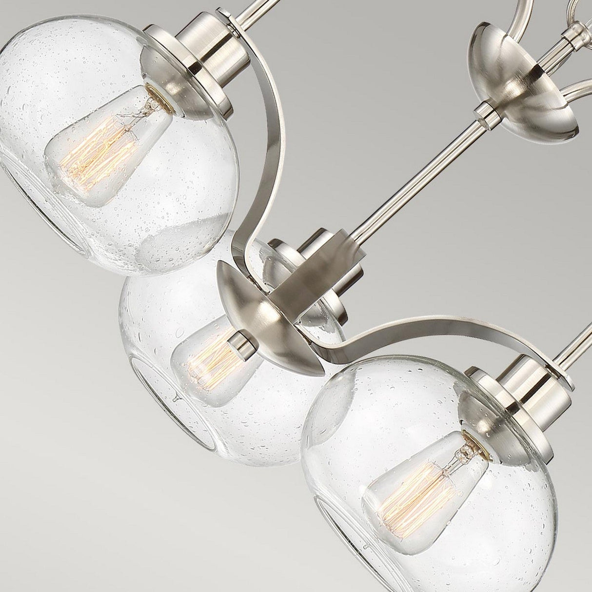 Introducing the Trilogy 3 Light Chandelier in Brushed Nickel: A close-up reveals three seeded glass globe lampshades, each housing a vintage-style filament bulb. The semi-flush fixture's brushed nickel finish offers a sleek and contemporary appearance against a neutral backdrop.