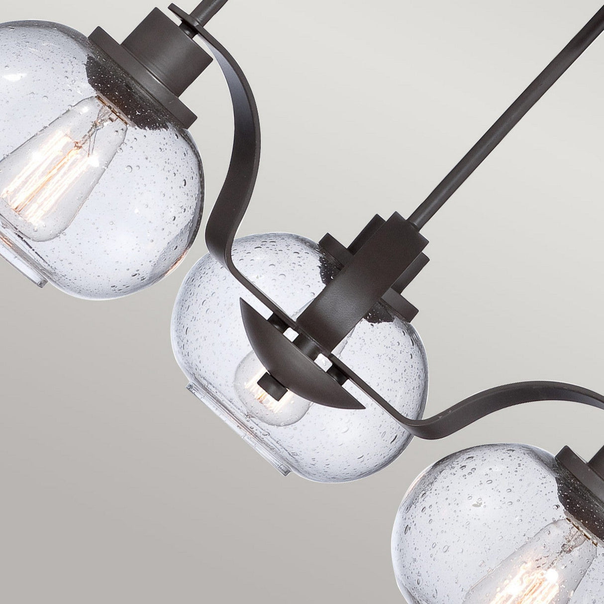 A close-up of the Trilogy 3 Light Chandelier - Old Bronze showcases three round seeded glass pendant lights with visible bulbs. The speckled texture complements the chandelier's old bronze metal hardware, set against a light gray background.