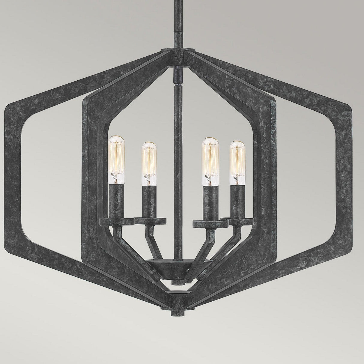 The Vanguard 4 Light Chandelier - Old Black showcases a modern hexagonal frame made from geometric black metal, featuring four exposed vertically aligned bulbs. Its industrial-inspired design stands out against a simple light gray background.