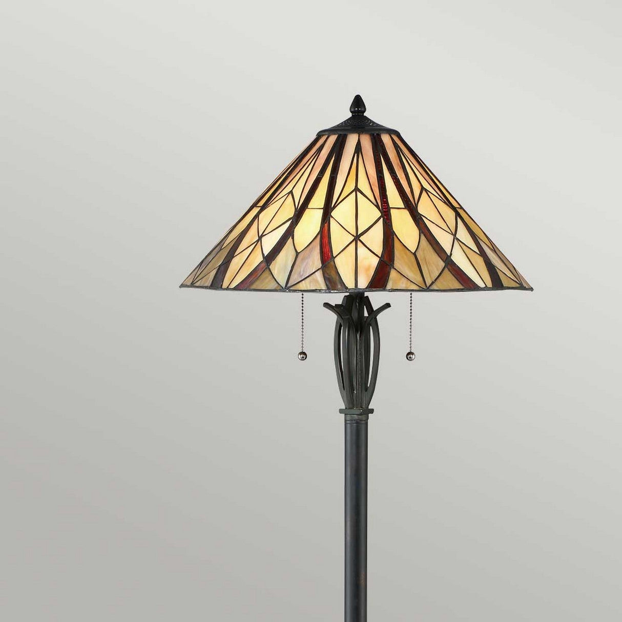 The Victory 2 Light Tiffany Floor Lamp in Valiant Bronze features a tall, elegant design with a stained glass shade that displays an intricate geometric pattern in warm hues of yellow, red, and beige. It includes two pull chains and stands against a light gray background, offering exquisite ambient lighting.