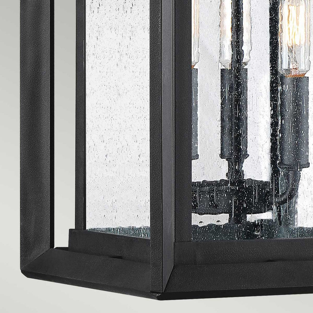The Wakefield Outdoor 3 Light Large Wall Lantern in Earth Black showcases a close-up of its black metal and glass design. This lantern-style light fixture includes three exposed bulbs and a textured glass surface, incorporating Arts and Crafts design elements within its contemporary geometric frame.