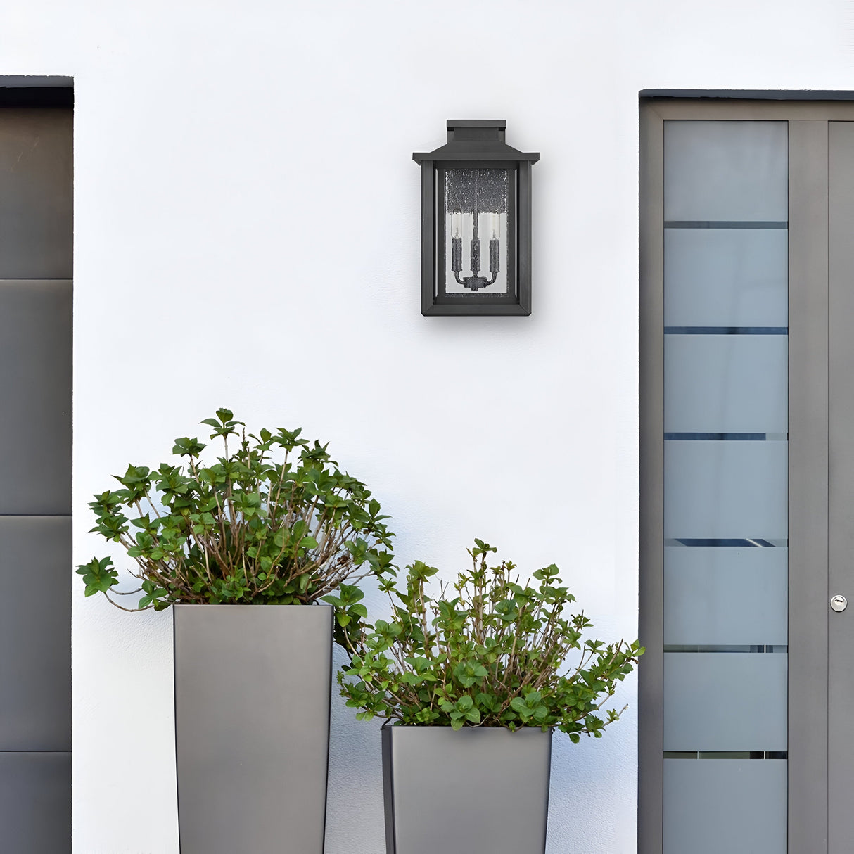 A modern exterior wall showcases a gray door adorned with horizontal glass panels. Below the Wakefield Outdoor 3 Light Large Wall Lantern in an Earth Black finish, two tall gray planters containing vibrant green plants are positioned. The white background wall enhances the minimalist and contemporary aesthetic inspired by Arts and Crafts design principles.