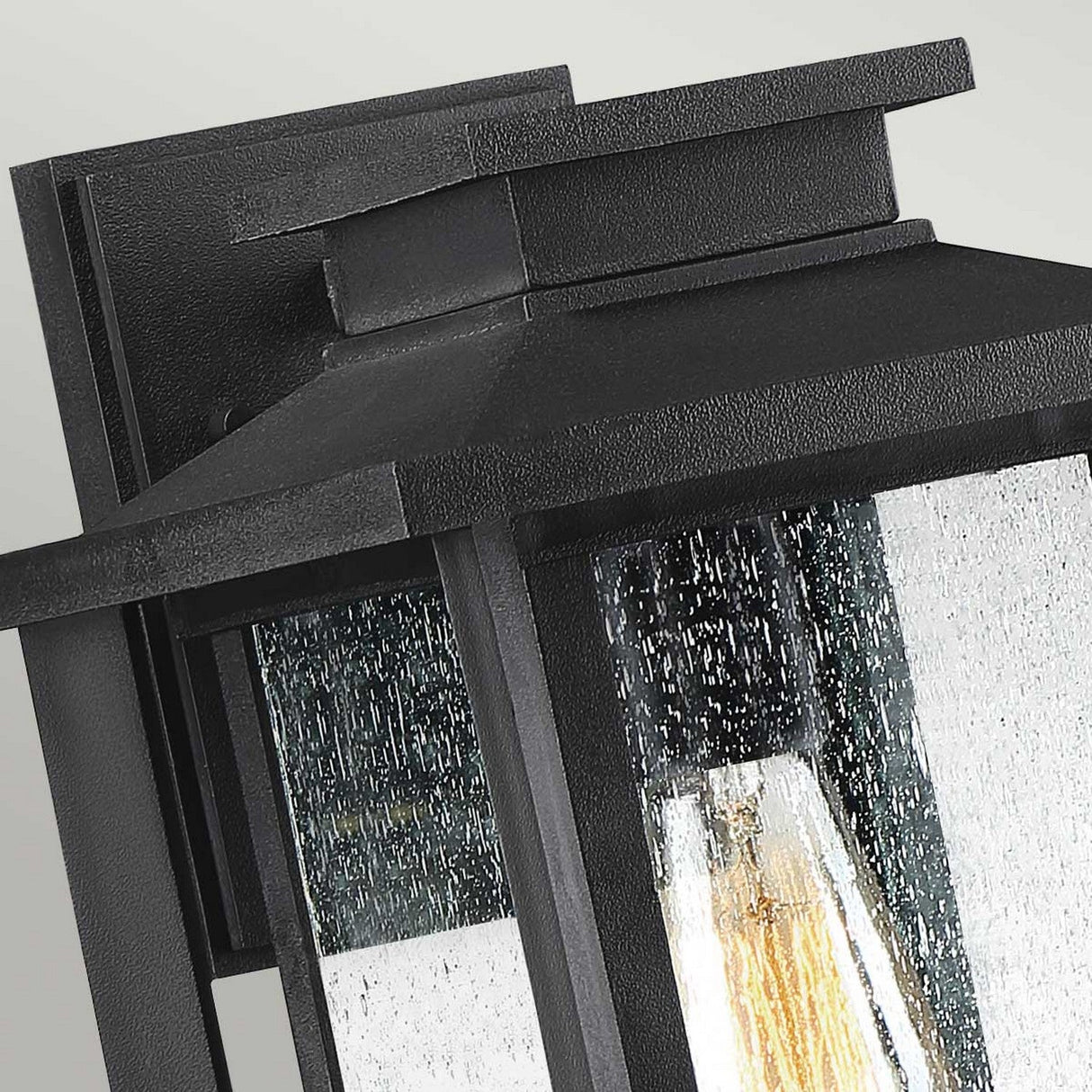 A detailed view of the Wakefield Outdoor 1 Light Medium Wall Lantern showcases its elegant Earth Black finish. This rectangular fixture, embellished with clear glass panels featuring a water-like texture, reveals a lightbulb inside. Installed on a light gray wall, this weather-resistant lantern brings sophistication to any outdoor area.