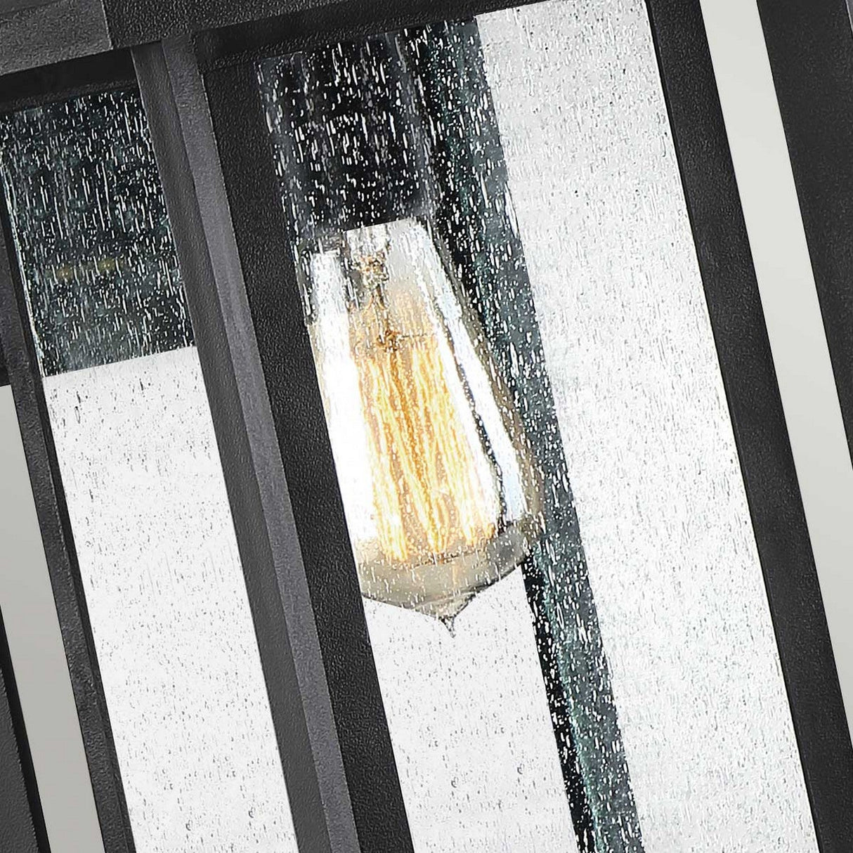 A close-up view of the Wakefield Outdoor 1 Light Medium Wall Lantern in Earth Black, featuring a vintage-style light bulb. The weather-resistant glass, adorned with rain droplets, adds texture and enhances its timeless charm.