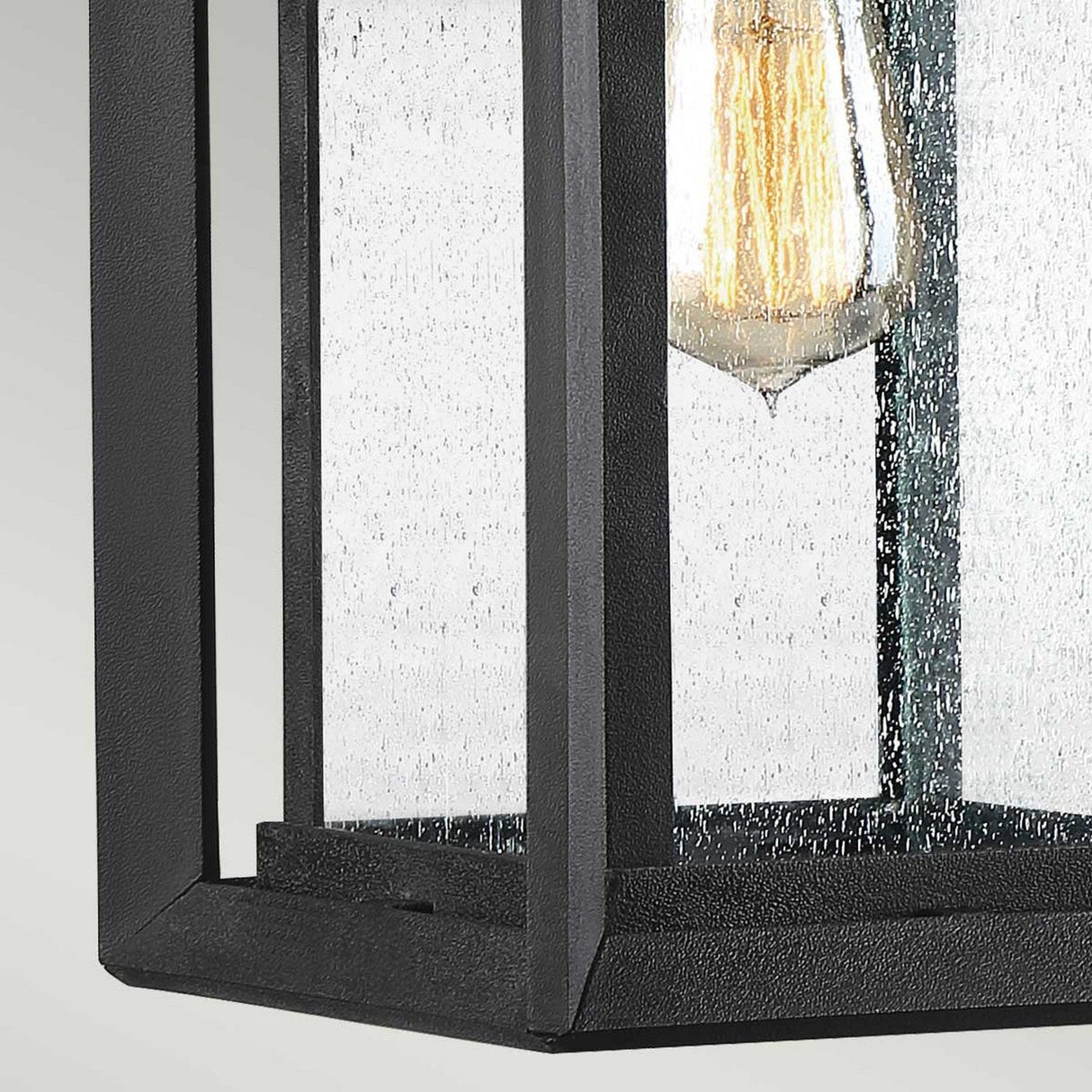 A close-up view reveals a rectangular black metal lantern featuring a glass panel with visible droplets. Inside, an exposed filament bulb radiates warm light. This Wakefield Outdoor 1 Light Medium Wall Lantern, finished in Earth Black, showcases a modern design that enhances the texture of its weather-resistant lighting.