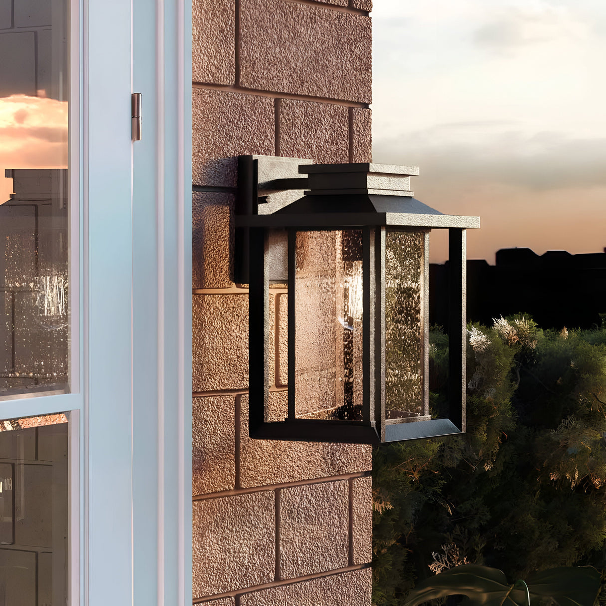 The Wakefield Outdoor 1 Light Medium Wall Lantern in Earth Black is installed on a brick wall, showcasing its modern design with clear glass. Its dusk illumination feature and weather-resistant qualities are complemented by a sunset's reflection in the nearby window, set against a backdrop of lush greenery.
