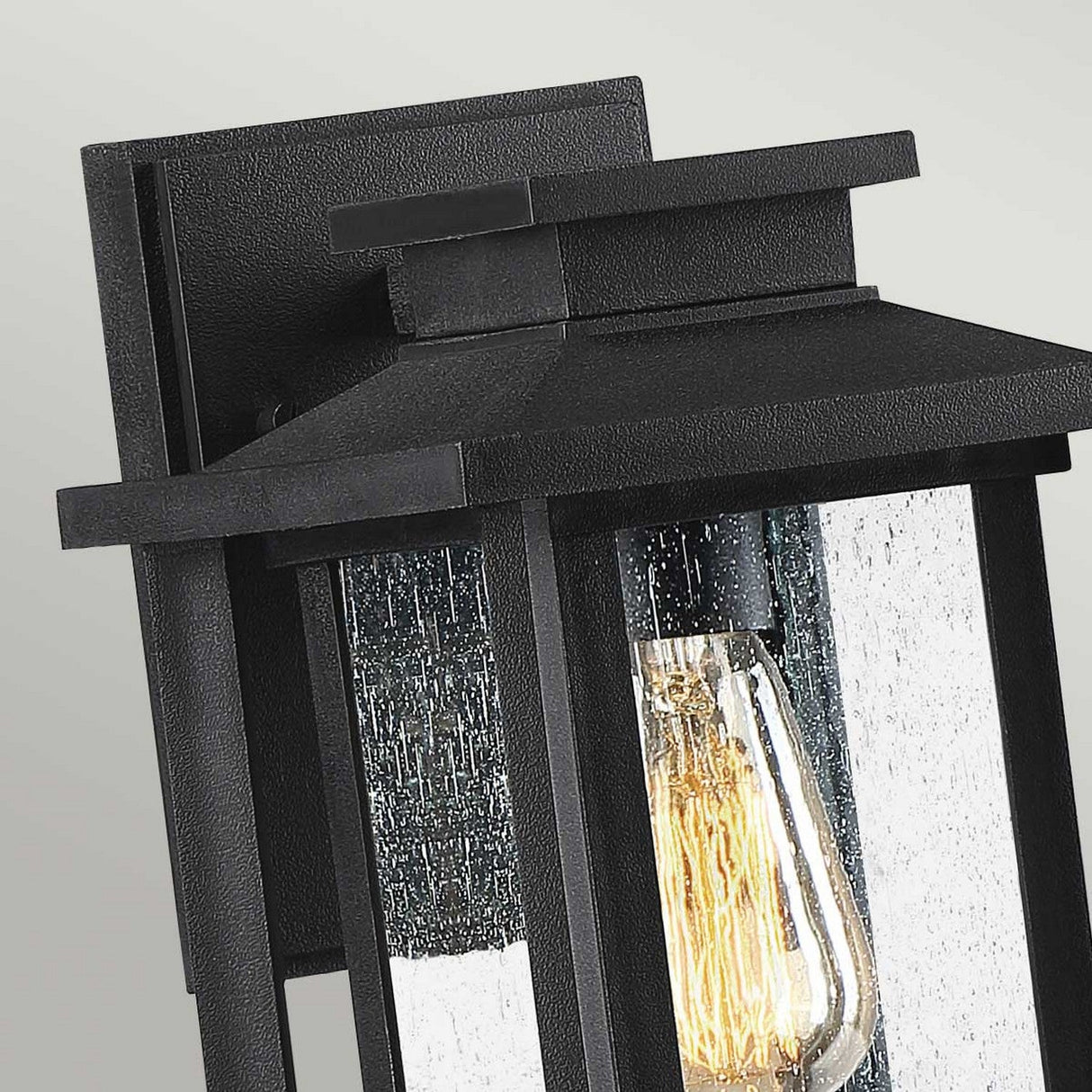 A detailed view of the Wakefield Outdoor 1 Light Small Wall Lantern in Earth Black showcases its rectangular design and clear glass panel. Inside, a vintage-style Edison bulb casts a warm glow. The lantern is firmly attached to a straightforward square backplate.