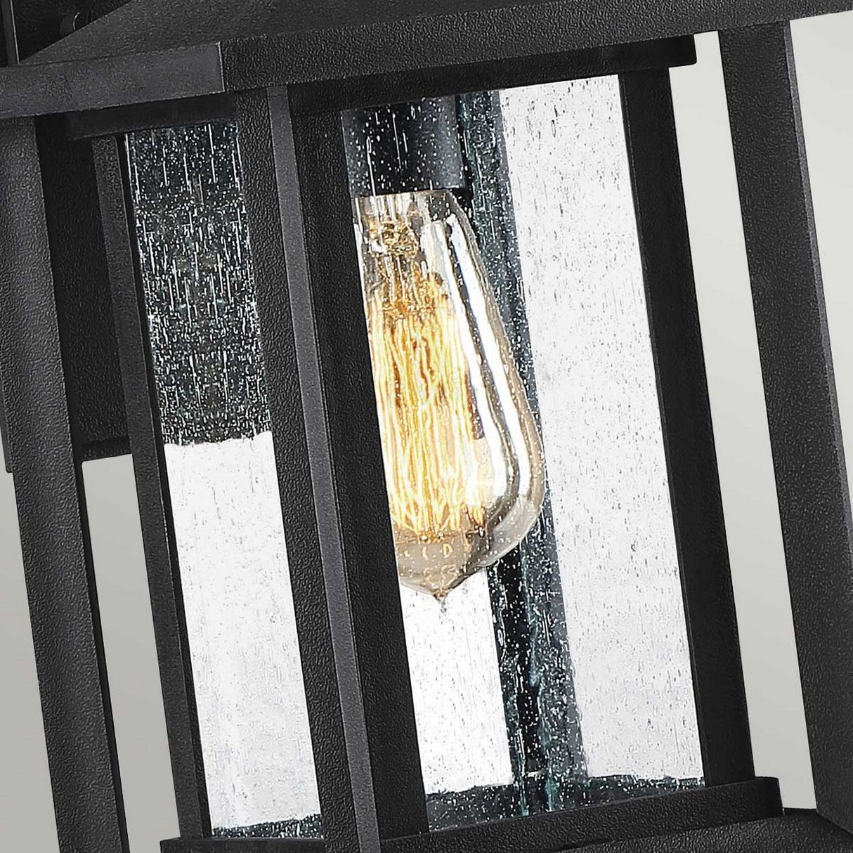 A close-up of the Wakefield Outdoor 1 Light Small Wall Lantern in Earth Black showcases its weather-resistant, vintage-style light bulb encased in a black metal and glass structure. The textured finish on the glass panels resembles droplets of water, while the bulb emits a warm glow, enhancing the lantern's elegant design.