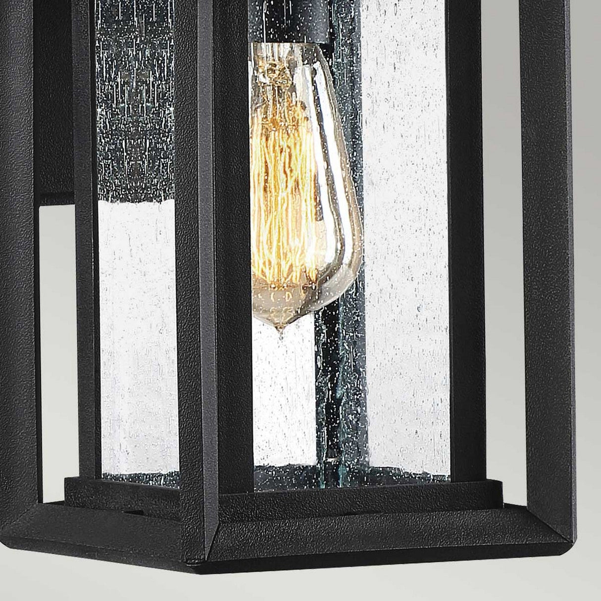 The Wakefield Outdoor 1 Light Small Wall Lantern in Earth Black features a close-up of its vintage-style black frame, with a clear glass enclosure to protect the weather-resistant Edison bulb inside. The bulb's visible filaments cast a warm glow against the textured glass surface, perfect for outdoor use.