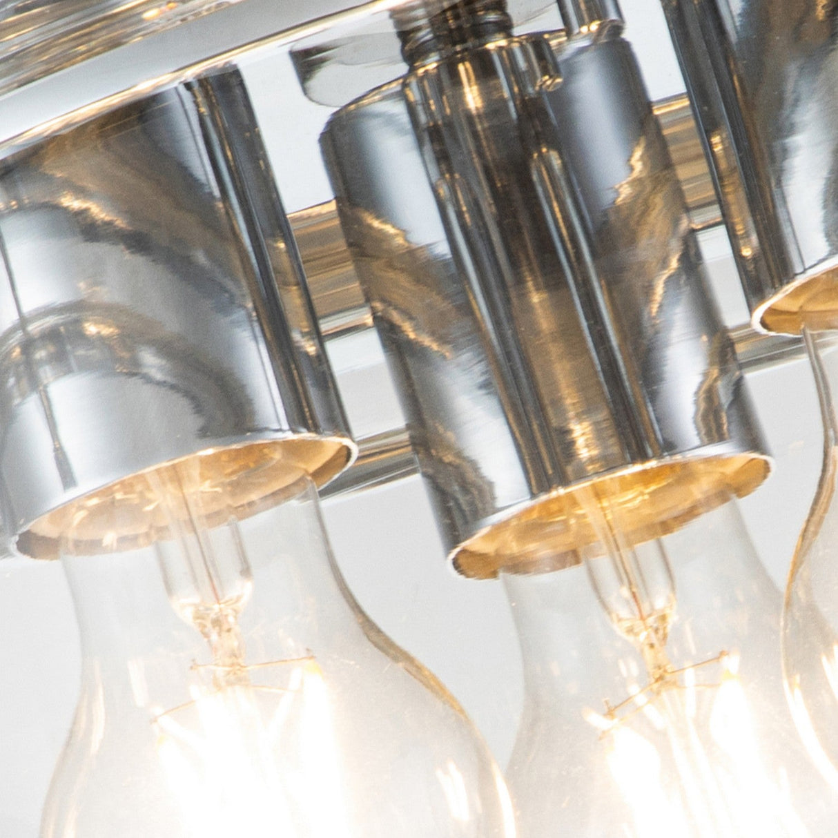 A close-up view of the Whistling 3 Light Flush fixture in polished chrome, featuring three glowing light bulbs in a metallic setting. Each bulb is lined up in a gleaming holder, emitting warm light that enhances the modern aesthetic of bathroom lighting.