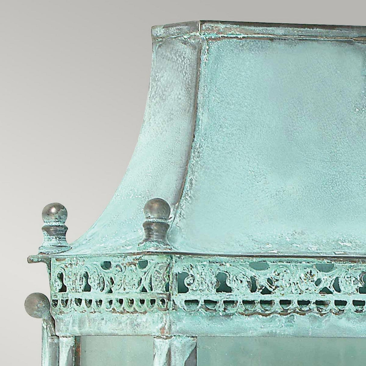 A detailed view of the Regents Park 1 Light Outdoor Wall Light - Verdigris showcases its vintage copper with a verdigris finish, highlighting a weathered patina. This outdoor wall light features intricate details and a curved top adorned with small finials at the corners, and its aged textures accentuate its classic lantern design.