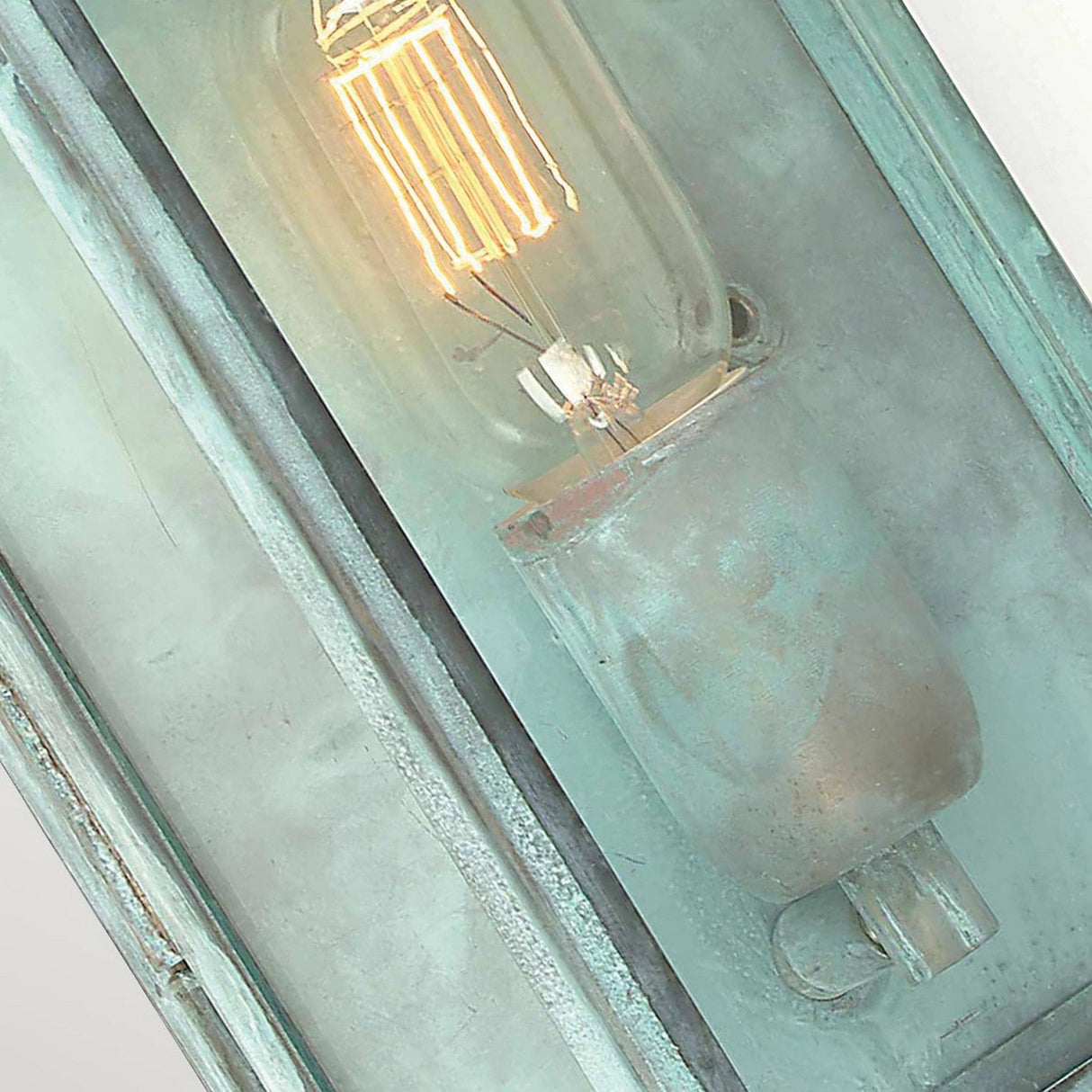 Close-up of the Regents Park 1 Light Outdoor Wall Light in a rustic, industrial style, featuring an exposed, glowing vintage Edison bulb. The lantern design is encased in a weathered metal frame with a verdigris finish, highlighting its textured surface and antique appearance.
