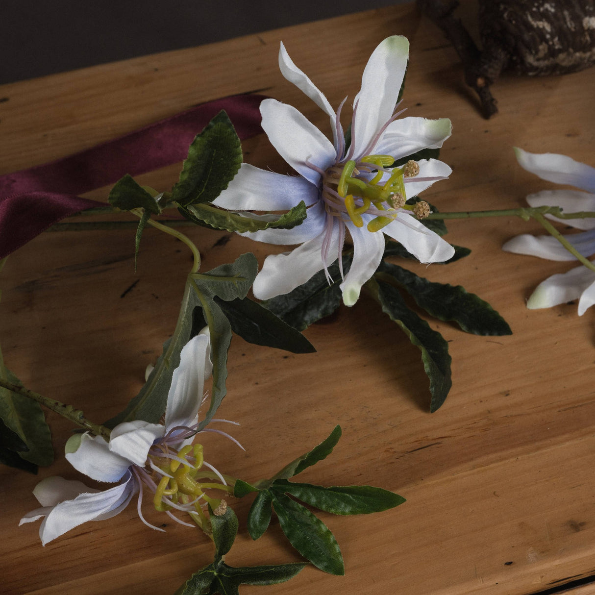 Hill Interiors Passion Flower Spray with white layered petals, fine filaments and green leaves, displayed on a wooden surface with burgundy ribbon.