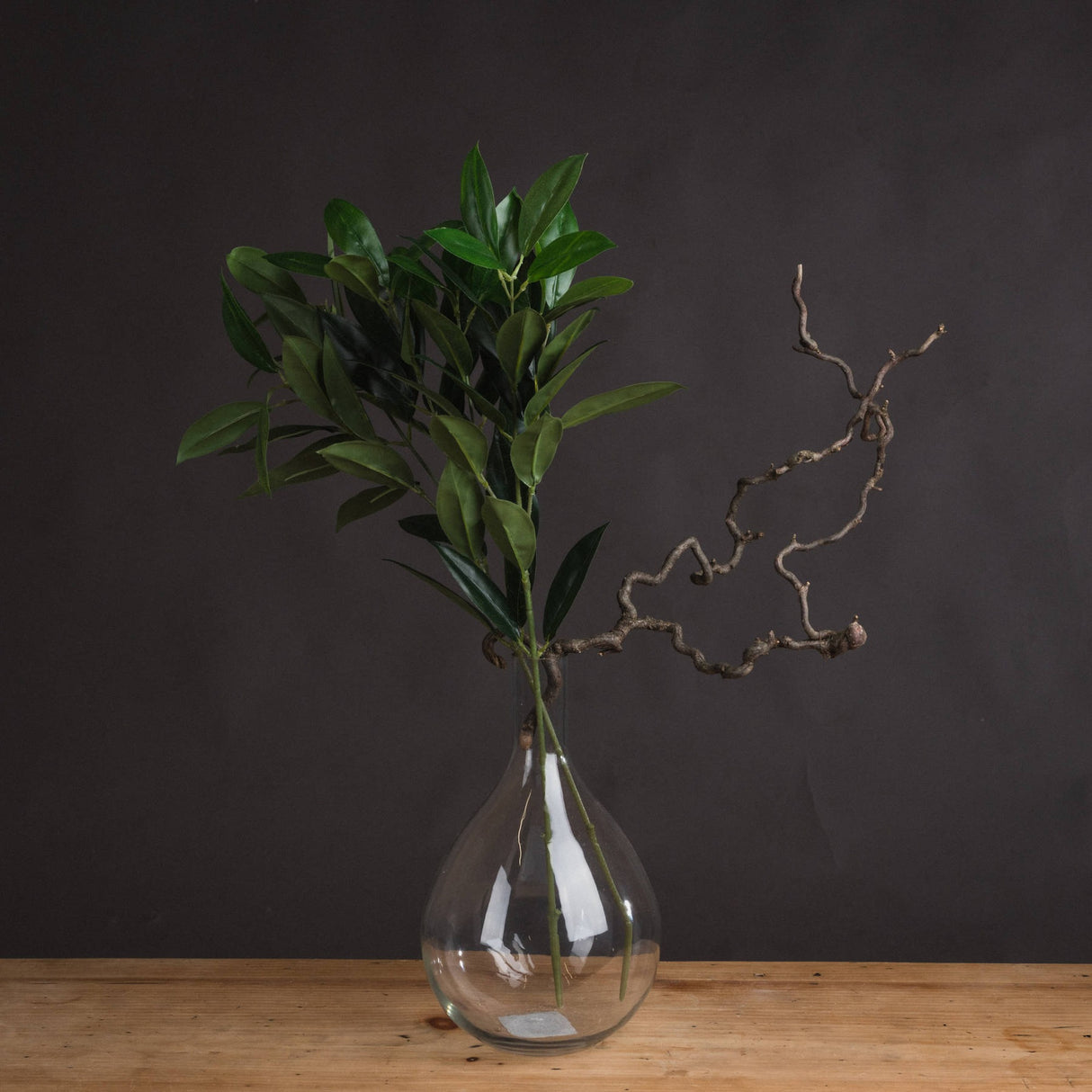 Hill Interiors Faux Bay Leaf Spray with tapered green leaves displayed in a clear glass bottle vase alongside a twisted branch on a wooden surface.