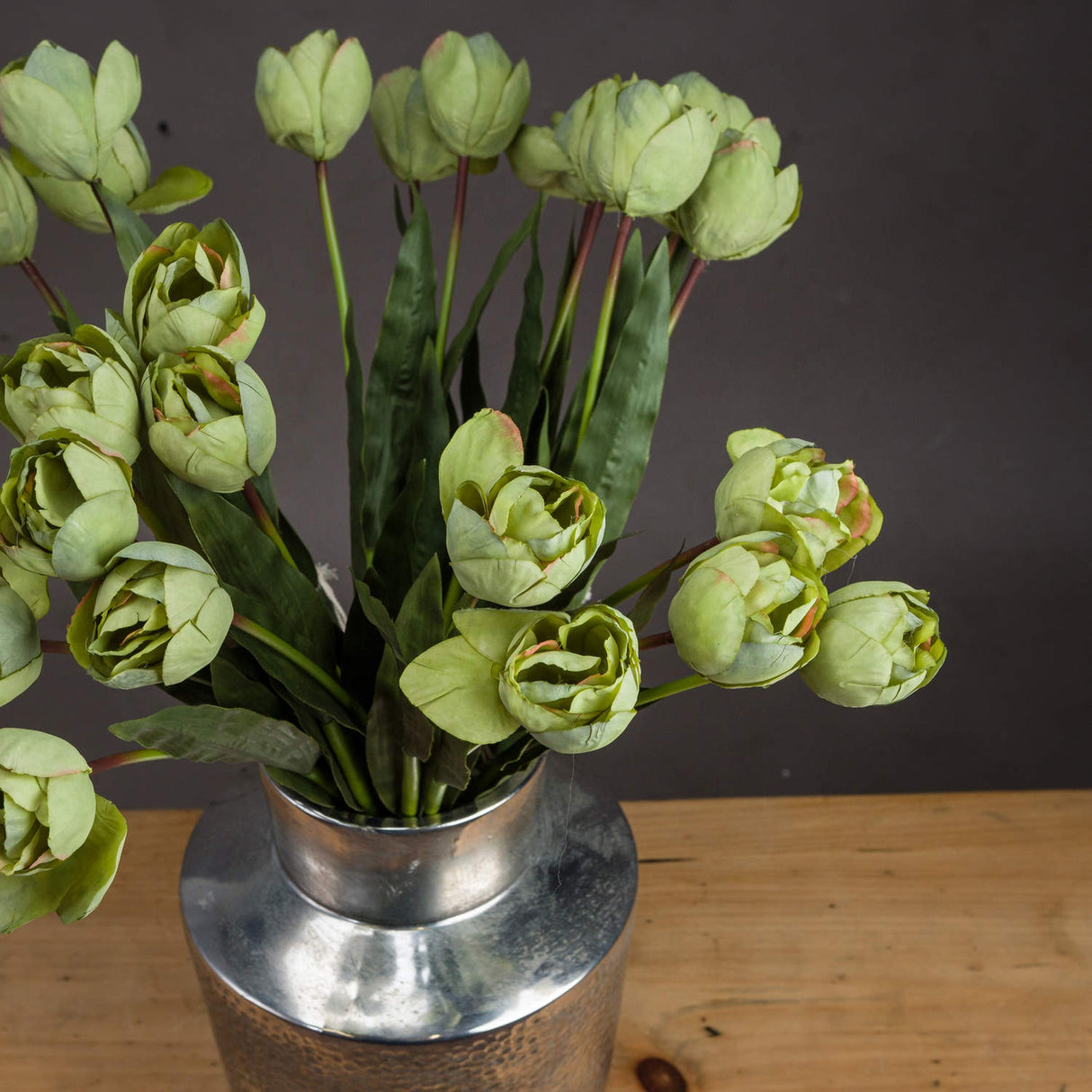 Hill Interiors Green Tulip faux flower stems with layered cupped heads and slim leaves, arranged in a hammered silver vase on a wooden surface.
