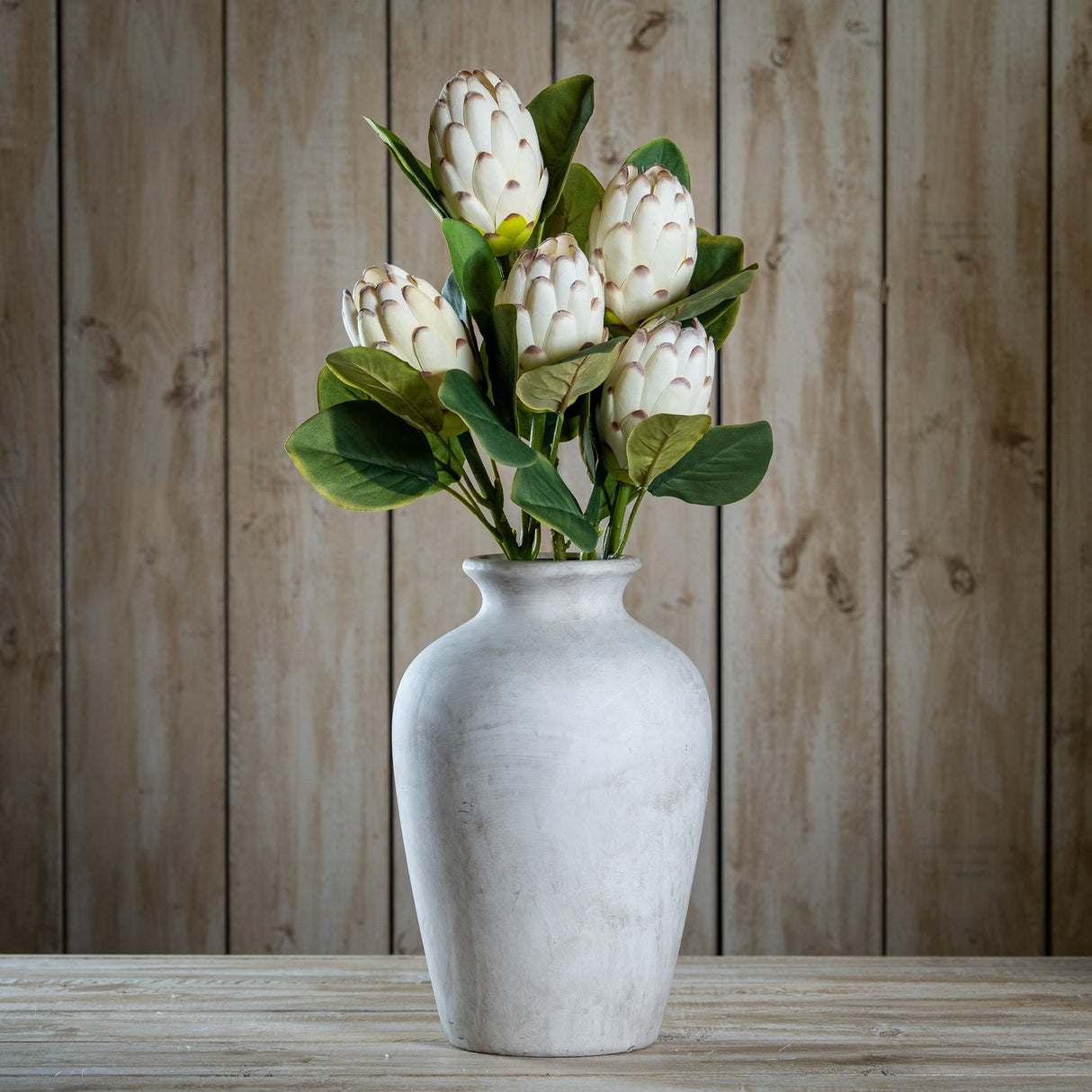 Hill Interiors Closed White Protea faux stems arranged in a distressed white ceramic vase on a wooden surface against a rustic timber plank backdrop.