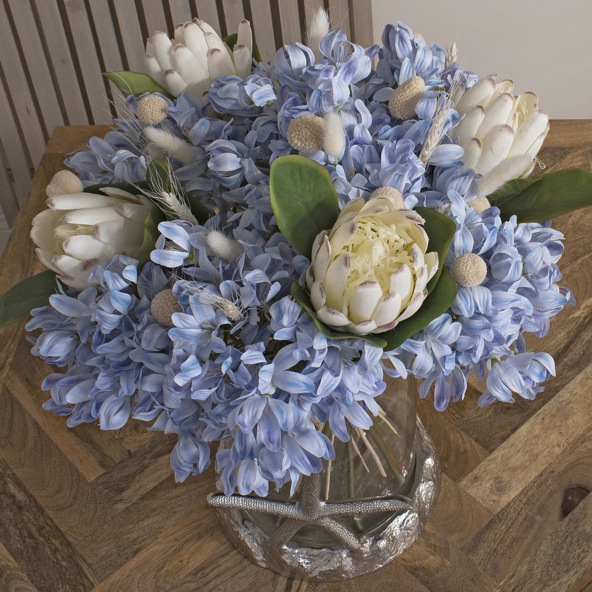 Hill Interiors Closed White Protea faux stems displayed with blue agapanthus and bunny tail grasses in a glass vase on a wooden table.