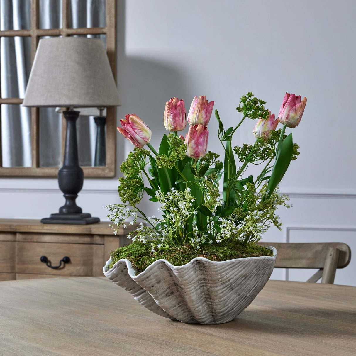 Hill Interiors white gypsophila spray with pink tulips and moss in a ridged clam-shaped bowl on a wooden dining table beside a lamp and mirror.