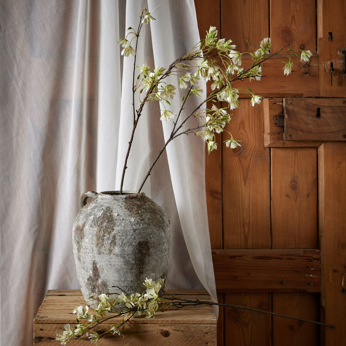 Hill Interiors Elaeocarpus Stem with delicate white blossoms and green leaves arranged in a rustic weathered stone vase beside draped linen and a wooden door.