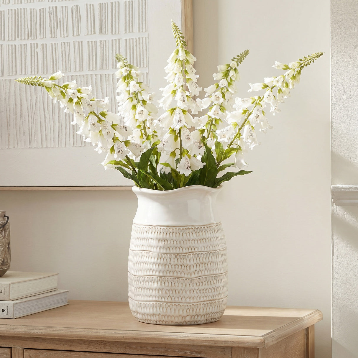 Hill Interiors White Foxglove faux flower stems with tall speckled bell-shaped blooms, displayed in a textured cream ceramic vase on a wooden console.