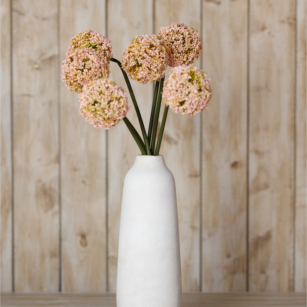 Hill Interiors Pink Allium Stems with pom-pom blooms and green stalks arranged in a tall white ceramic vase against a light wood plank backdrop.