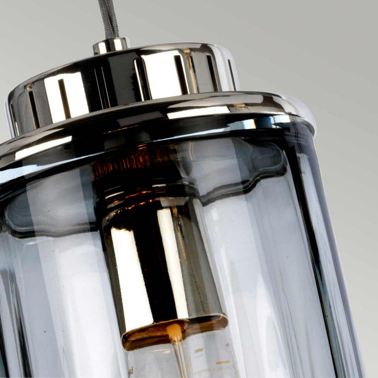 A close-up of the Reno 1 Light Pendant featuring a polished nickel top and smoke glass casing, showcasing the bulb inside. The pendant light is suspended by a cord against a plain background.