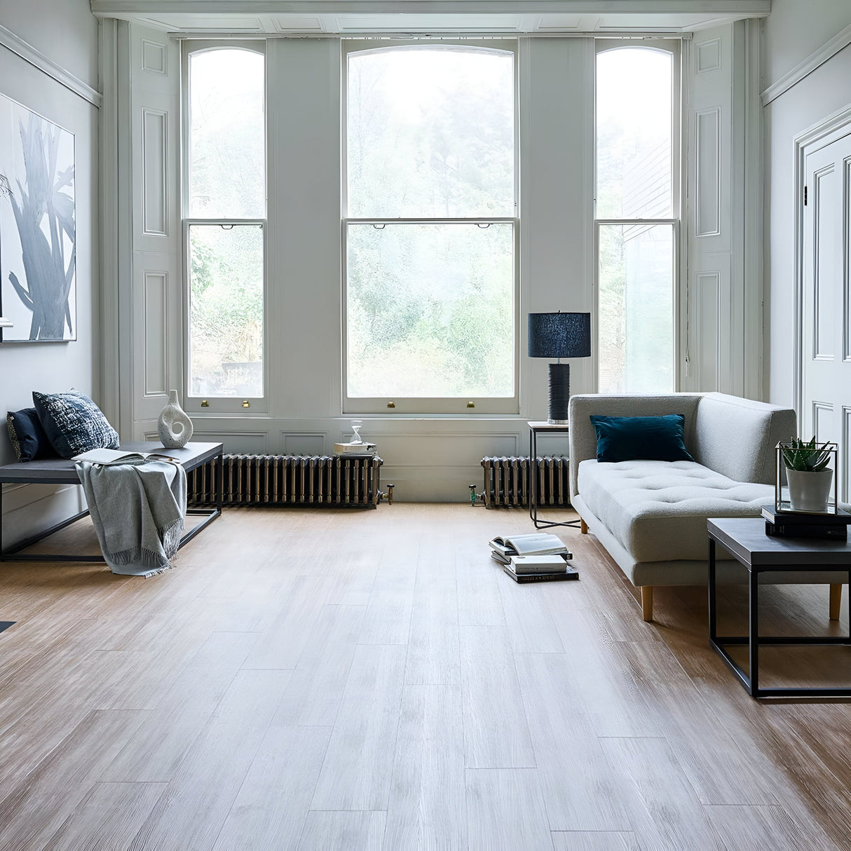 A bright, modern living room with large windows allows natural light to fill the space. It features a gray sofa and a Ripple 1 Light Table Lamp - Black With White Shade that offers contrast decor on the coffee table, accompanied by books and a plant. A painting adorns the wall, while the light wood floor enhances the spacious and minimalistic feel.