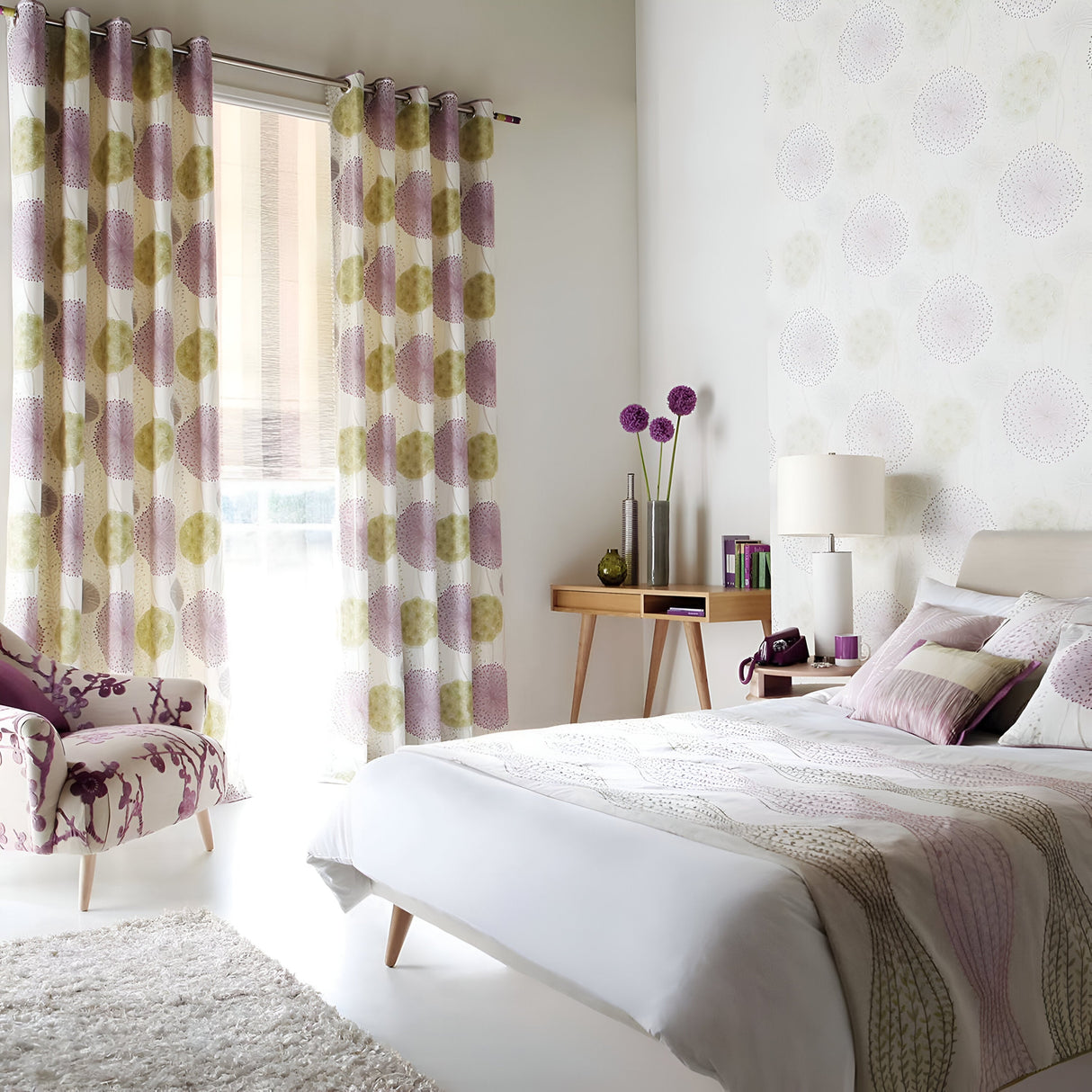 A modern bedroom with a minimalist design showcases a white and pastel color palette. The room includes a bed with crisp white linens, a floral chair, and curtains featuring large circles. A wooden desk decorated with accents is complemented by the Ripple 1 Light Table Lamp - White With White Shade, along with decorative pillows and eye-catching wall patterns.