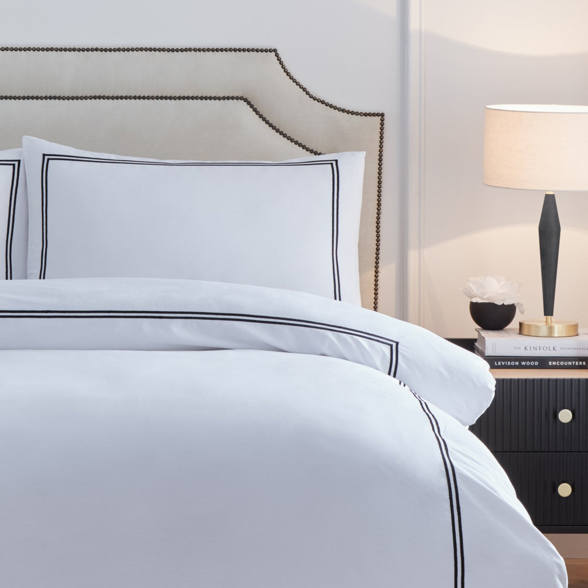 Elara white cotton duvet cover set with black twin-line embroidered border, styled on a bed with matching pillowcases beside an upholstered headboard and nightstand.