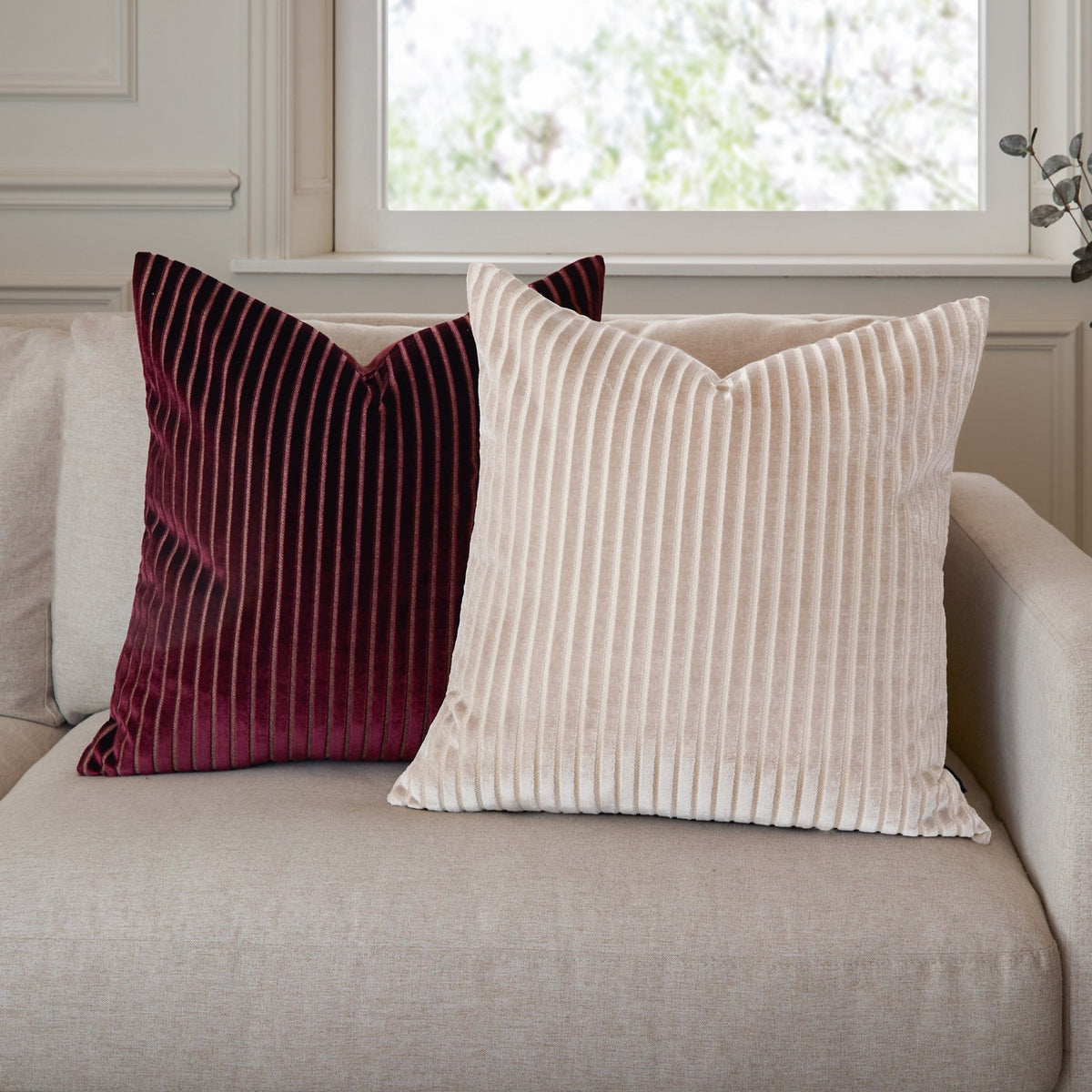 Savoy Stripe Polyester Velvet Cushion in Oyster, a square cushion with ribbed cream velvet front, shown on a beige sofa beside a burgundy striped velvet cushion.