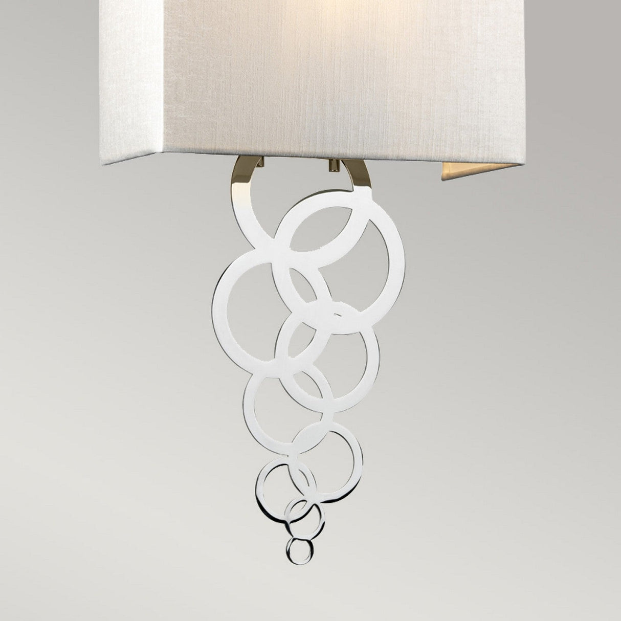 The Rosa Medium 1 Light Wall Light in polished chrome showcases a modern design with a rectangular fabric shade and a decorative hanging accent of intersecting metallic circles, offering a stylish and elegant look.