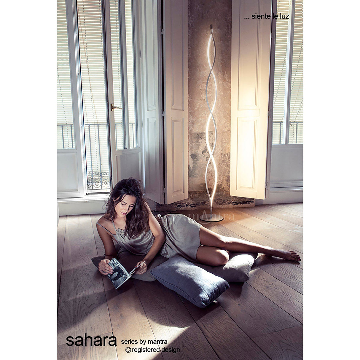 A woman in a light dress lounges on cushions, reading near tall shuttered windows. Behind her, the Sahara LED Floor Lamp 21W 3000K in Silver & Chrome casts gentle ambient lighting across the wooden floor.