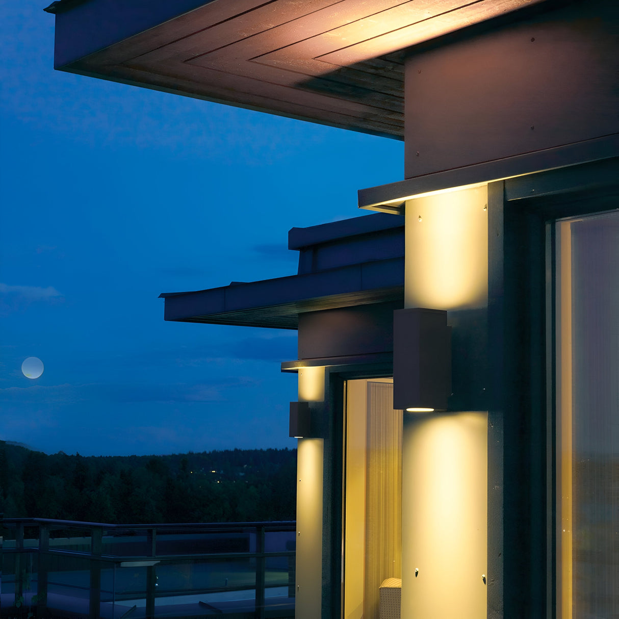 At dusk, a contemporary home exterior showcases warmly illuminated Sandvik Outdoor 2 Light Up & Down Wall Lights in a sleek graphite finish, rated IP65. The sky is a deep blue, and a full moon glows above the tranquil landscape of trees.