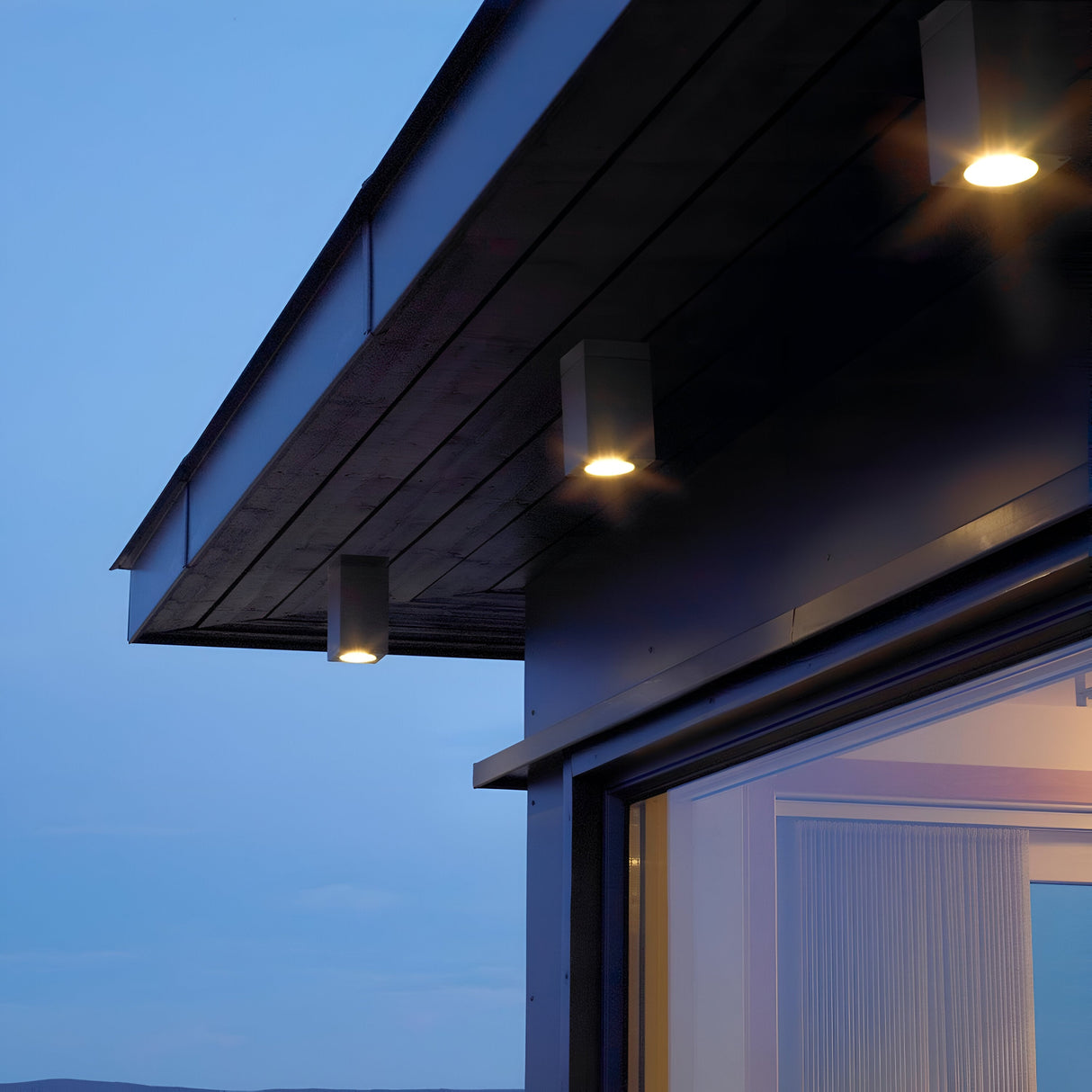 At dusk, a contemporary building with a sloped roof highlights the Sandvik Outdoor 1 Light Down Ceiling Light in Graphite, boasting an IP65 rating. The gentle illumination accentuates the graphite finish of the structure against the clear evening sky. A partially open sliding glass door is visible, revealing an interior with closed curtains.