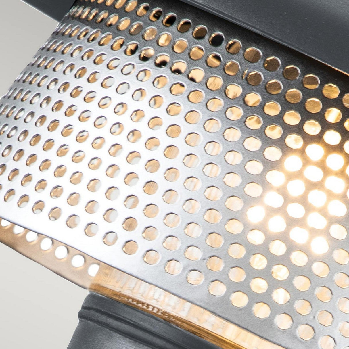 A close-up view of the Saturne 1 Light Outdoor Wall Light - Grey reveals circular perforations in its metal surface, reminiscent of a silver mesh diffuser that gently diffuses light. The texture and arrangement of these openings produce an intriguing visual effect, much like those seen in a modern outdoor wall light design. The background is softly illuminated.