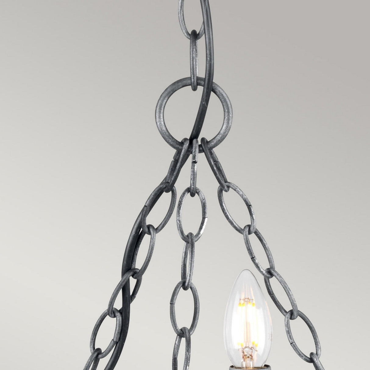The Saxon 3 Light Chandelier in Black/Silver Patina presents a close-up view, showcasing its medieval charm with exposed bulbs. It features metal chains in a looped design hanging from the ceiling. Its illuminated bulbs provide warm lighting, seamlessly combining modern elegance against a neutral gray background.
