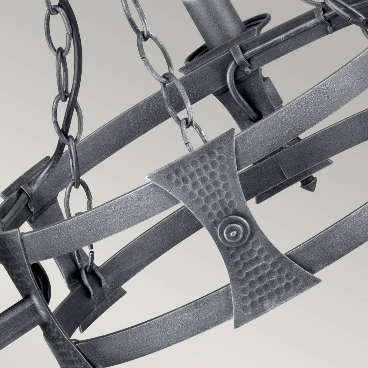 A close-up of the Saxon 3 Light Chandelier in Black/Silver Patina reveals a metallic spherical design made up of interlocking iron rings and chains, radiating a medieval charm. The surface is textured with small circular indentations, showcasing intricate craftsmanship similar to that of a chandelier. It stands against a plain, light gray background.