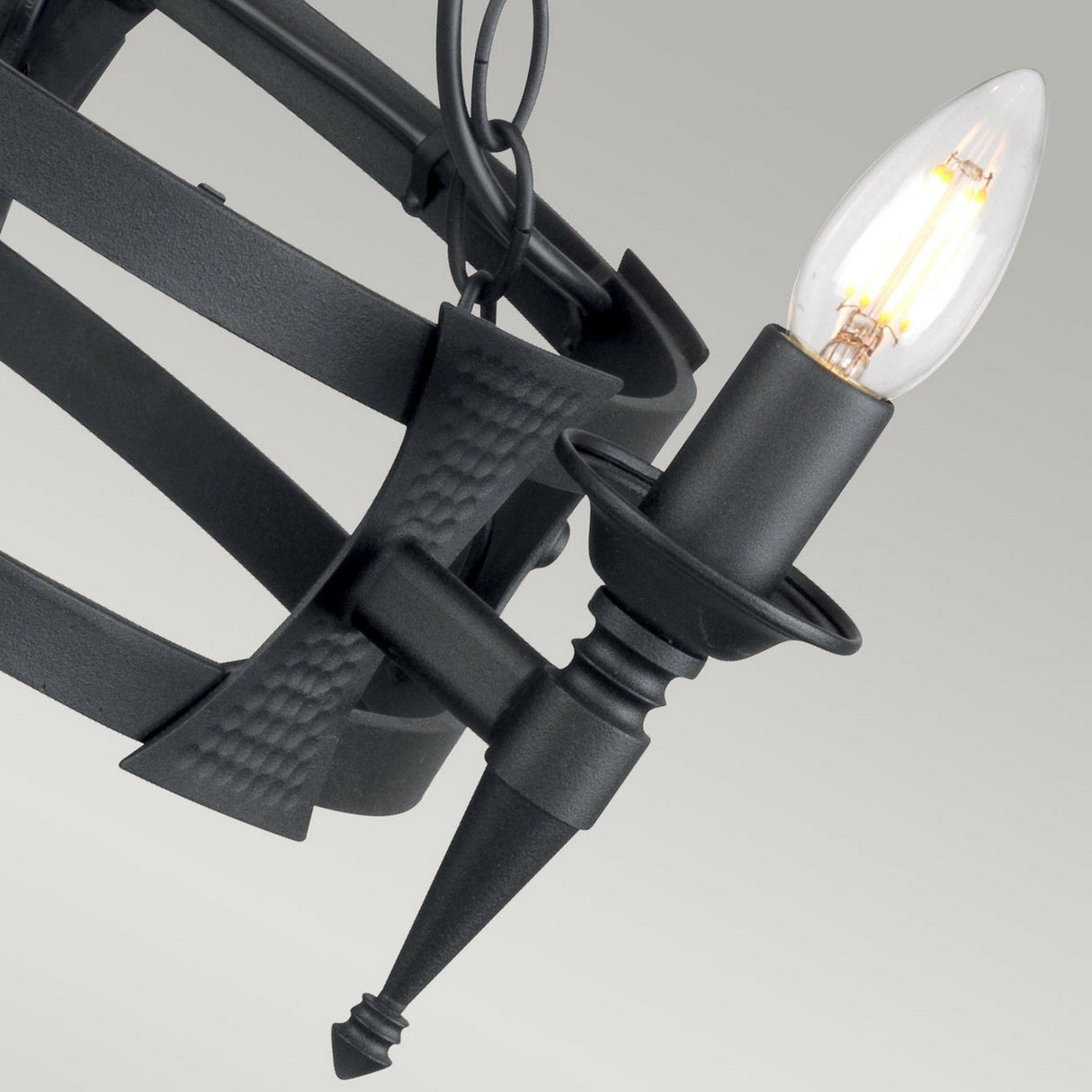 Close-up of the Saxon 3 Light Chandelier - Black, featuring a single lit candle-shaped bulb. The wrought iron design highlights a hammered texture and curved lines, presenting an elegant and modern appearance against a gray background.