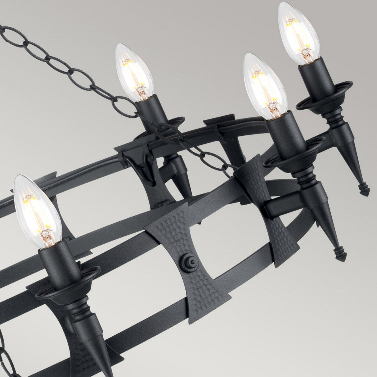 The Saxon 8 Light Chandelier - Black showcases a medieval design with an elegant circular frame, featuring five candle-shaped bulbs. It hangs from a chain and casts a warm glow that creates an inviting ambiance against a light gray background.